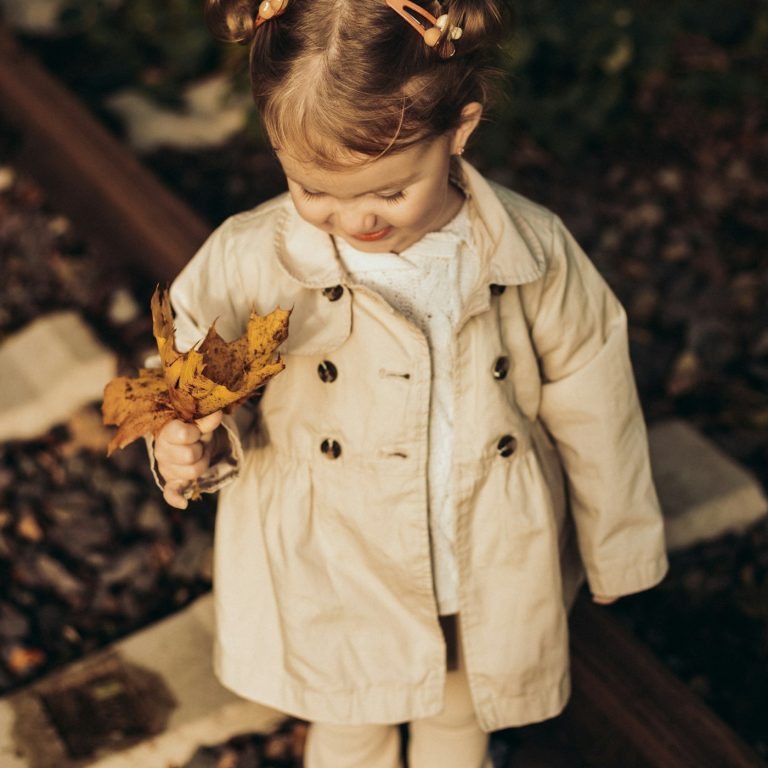 A toddler in a beige trench coat holding autumn leaves